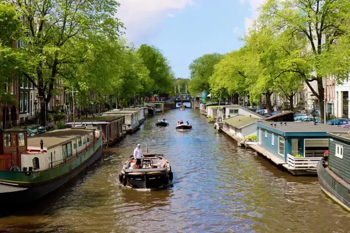 small open canal tour boat on an Amsterdam canal with house boats on both sides and other boats behind it