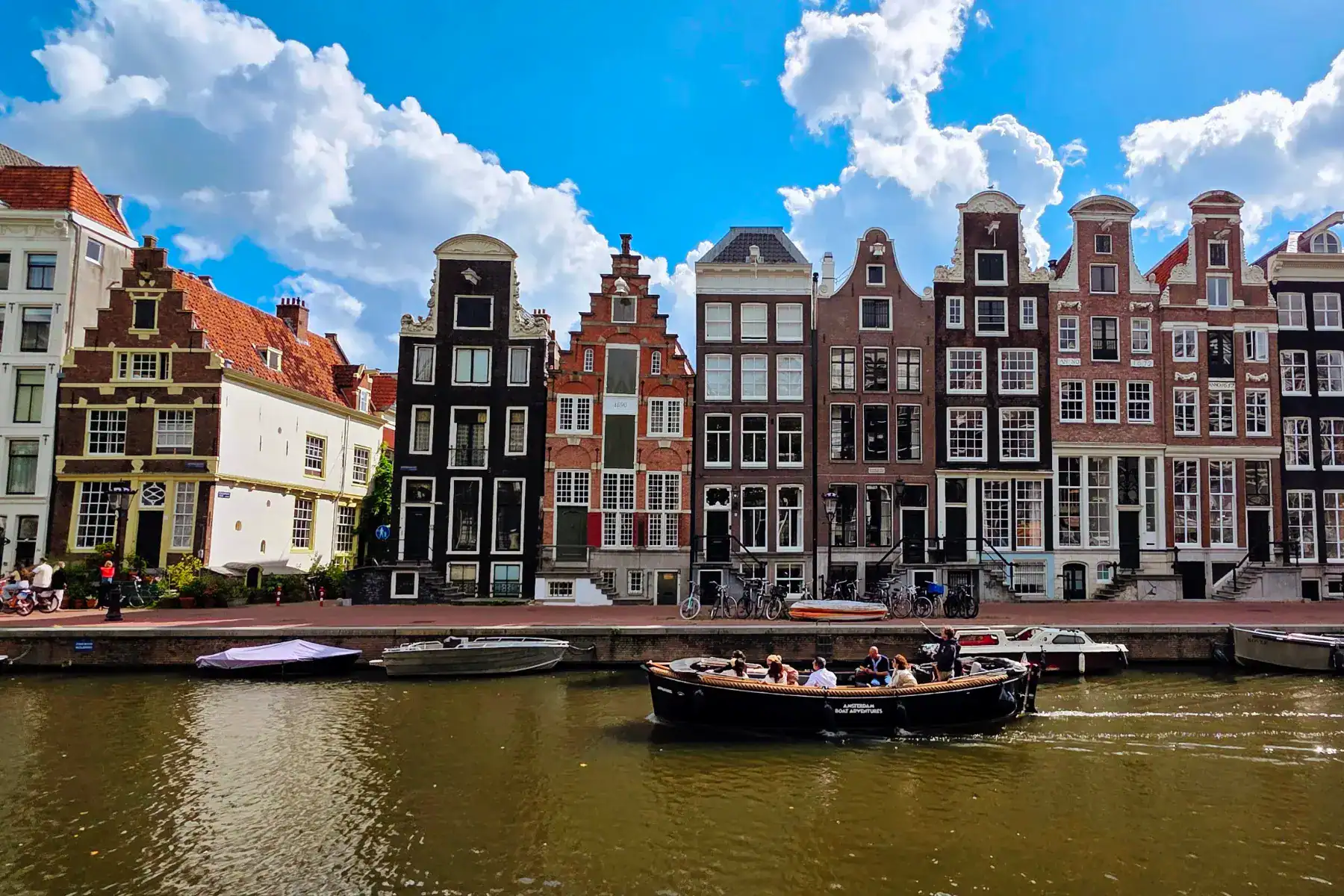 Colorful historic buildings along a canal in Amsterdam, with a boat cruising under a bright blue sky.