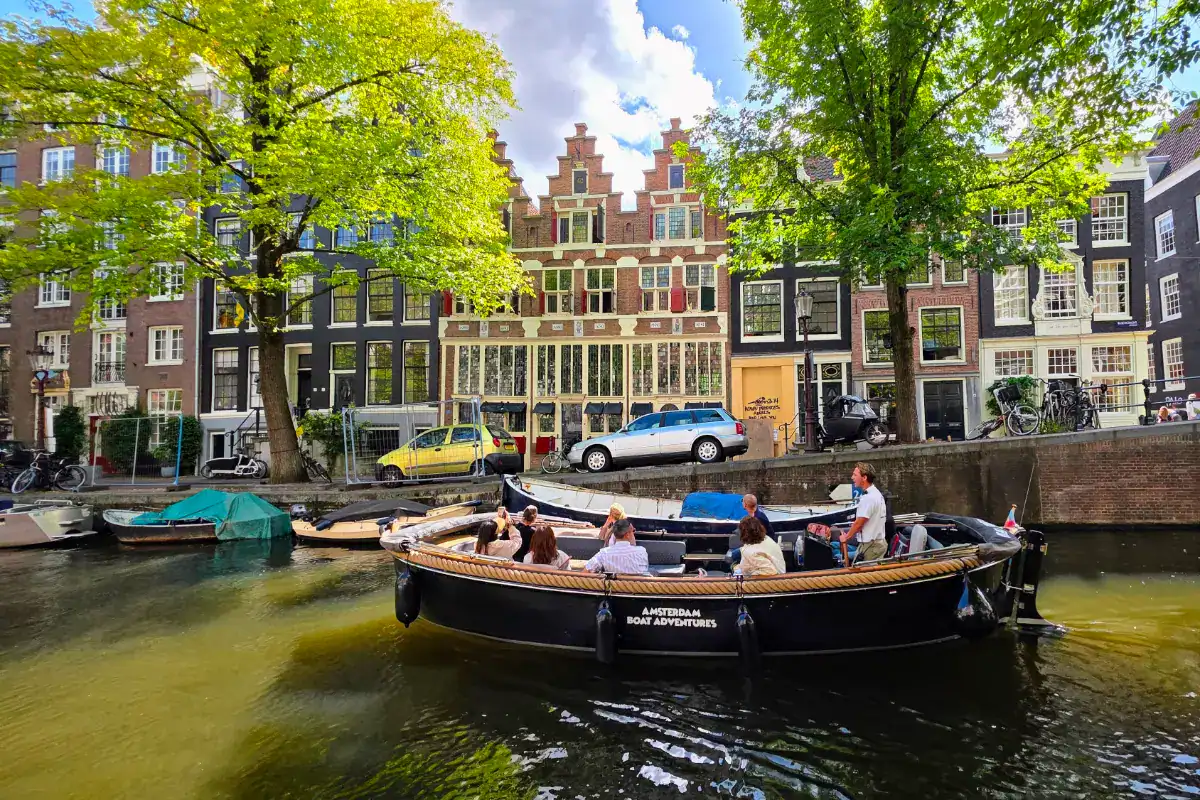 Canal tour boat in Amsterdam with historic buildings and lush trees along the water.