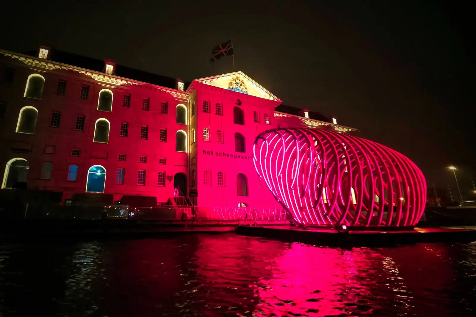 Night view of Amsterdam's illuminated historic building and modern art installation on the canal.